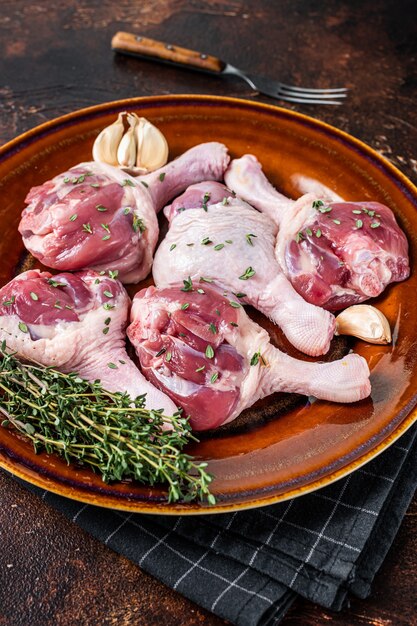 Viande de volaille crue - pilons de cuisses de canard sur une assiette rustique aux herbes. Fond sombre. Vue de dessus.