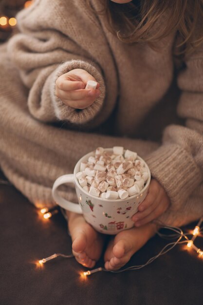 Vibes confortables verticales La main de l'enfant tient une tasse de chocolat chaud de Noël et mange de la guimauve dans un pull beige