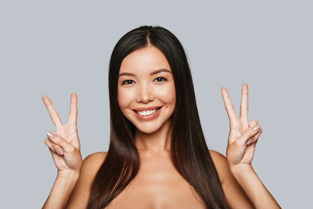 La victoire. Belle jeune femme asiatique gesticulant et souriant en se tenant debout sur fond gris