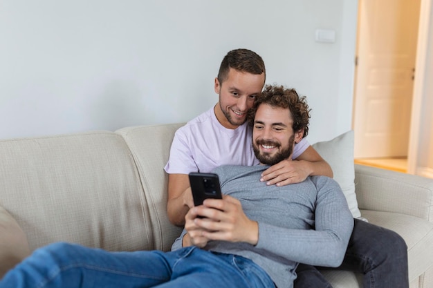 Vidéo d'un jeune couple gay mignon appelant leurs amis dans leur salon à la maison Deux amants souriants souriant joyeusement tout en saluant leurs amis sur un smartphone Jeune couple gay assis ensemble