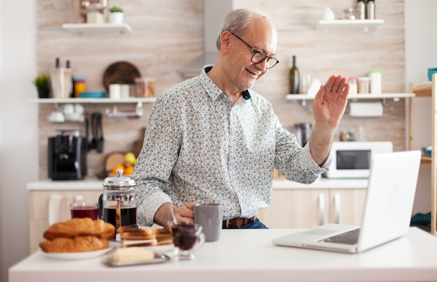 Vie quotidienne d'un homme âgé dans la cuisine pendant le petit-déjeuner à l'aide d'un ordinateur portable tenant une tasse de café, parlant en ligne avec des neveux saluant la webcam. Personne retraitée travaillant à domicile lors d'une réunion virtuelle, d'une conférence
