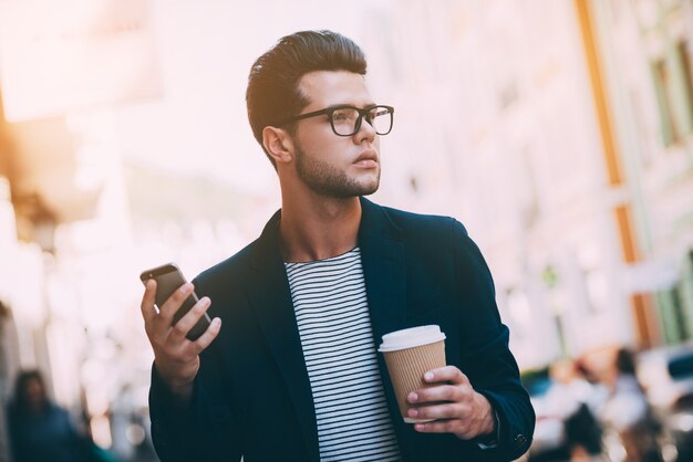 La vie en ville. Beau jeune homme en vêtements décontractés intelligents marchant le long de la rue tout en tenant une tasse de café et un téléphone intelligent