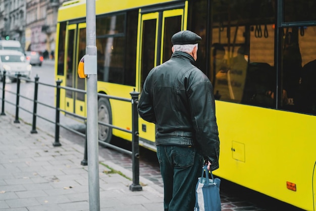 Vieil homme aux cheveux gris en casquette noire, veste en cuir noir et jeans restant près du grand bus jaune de la ville. Arrêt de bus. En attendant. Transport. Transport public