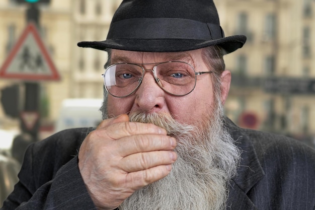 Vieil homme barbu couvrant la bouche avec sa main de près. Arrière-plan flou.