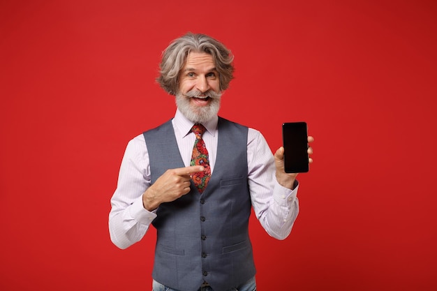 Vieil homme barbu moustache aux cheveux gris en chemise gilet cravate posant isolé sur fond rouge. Concept de style de vie des gens. Maquette de l'espace de copie. Doigt pointé sur le téléphone mobile avec écran vide vide.