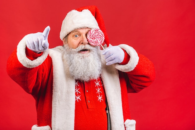 Vieil homme en costume de père Noël tenant un bonbon sucette isolé sur fond rouge