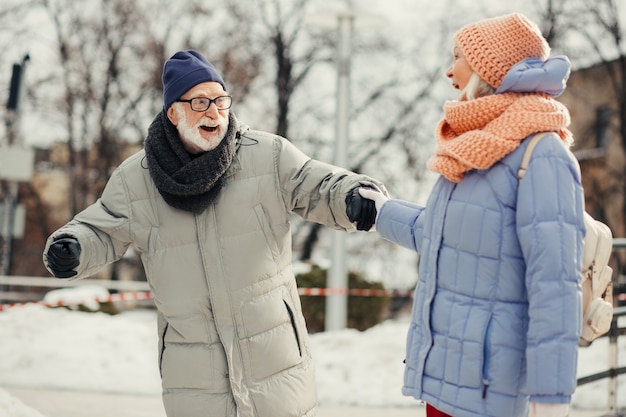 Un vieil homme émotif étant sur la patinoire avec sa femme bien-aimée et souriant tout en se tenant en équilibre sur la glace et en lui tenant la main