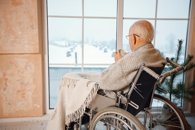 Vieil homme en fauteuil roulant assis près d'une grande fenêtre et buvant un thé