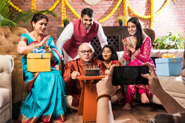 Vieil homme indien avec la famille célébrant l'anniversaire en soufflant des bougies sur le gâteau tout en portant des vêtements ethniques