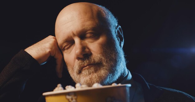 Un vieil homme regarde un film ennuyeux au cinéma et mange du pop-corn. Il dort les yeux fermés.