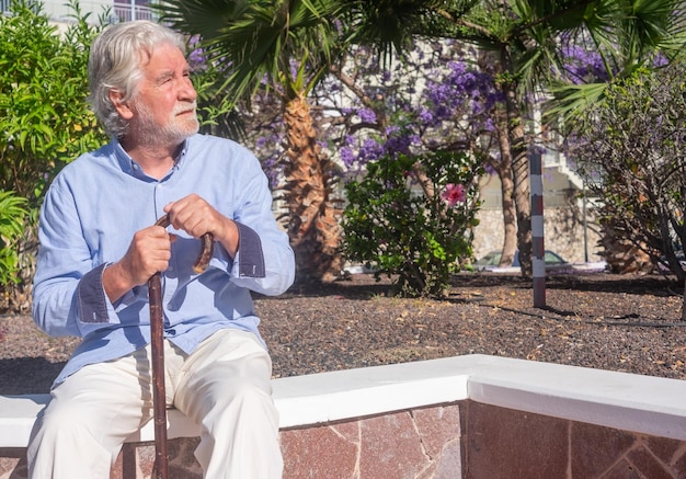 Vieil homme senior avec les mains sur la canne assis dans un parc public avec une expression triste Retraité âgé souffrant de maux de dos à l'aide d'un bâton de marche Arbre et fleurs sur fond