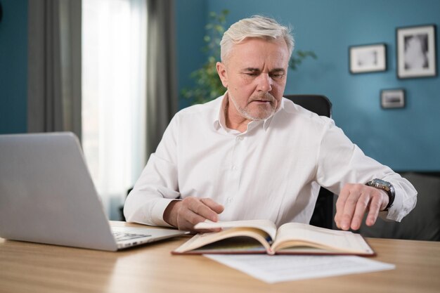 Vieil homme senior vérifiant les finances et les factures de la maison, il calcule les coûts à l'aide d'un ordinateur et écrit n