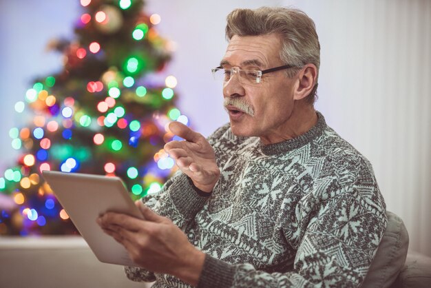 Le vieil homme avec une tablette passe un appel vidéo près du sapin de Noël