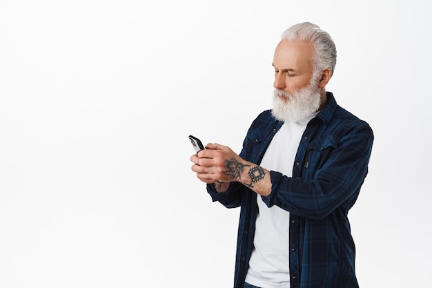 Un vieil homme avec des tatouages et des messages de barbe, lisant l'écran d'un téléphone portable avec désinvolture, utilisant un téléphone portable, debout sur un mur blanc.