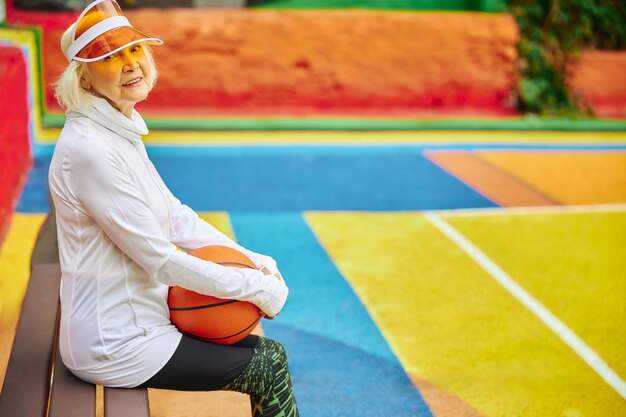 Vieille dame en bonne santé et joyeuse avec ballon de basket sur une place colorée à l'extérieur