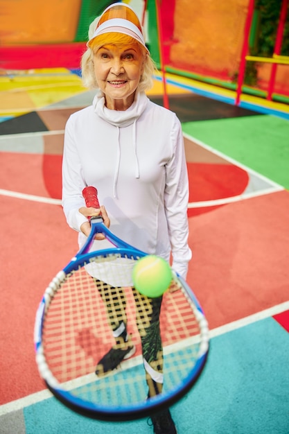 Vieille dame en bonne santé et joyeuse sur une place colorée à l'extérieur et tenant une raquette de tennis avec une balle