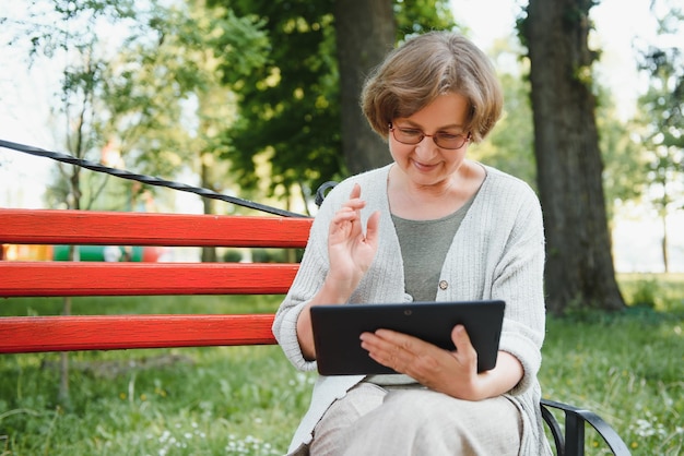Vieille dame confiante positive posant avec une tablette dans le parc Femme âgée aux cheveux gris en tenue décontractée assise sur un banc de parc et utilisant une tablette Concept de connexion sans fil