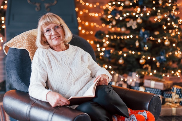 Vieille femme assise sur la chaise avec livre dans la salle festive du nouvel an avec des décorations de Noël.