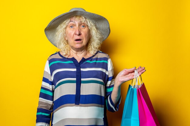 Vieille femme au chapeau tenant des sacs à provisions avec des achats sur fond jaune.