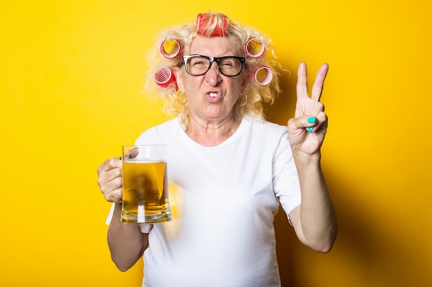 Vieille femme avec des bigoudis avec un verre de bière montre un geste de deux doigts, victoires et paix sur une surface jaune