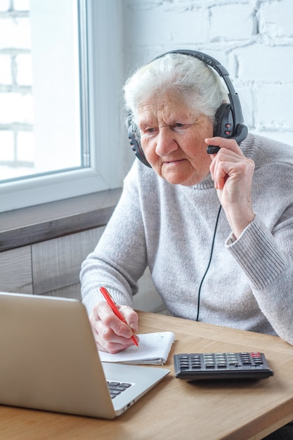 Une vieille femme est assise à une table devant un ordinateur portable avec un casque et écrit dans un cahier.