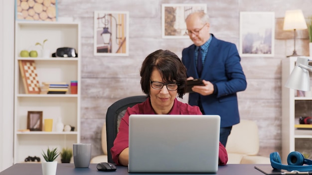 Une vieille femme étonnée après avoir lu une nouvelle sur un ordinateur portable dans le salon. Vieil homme utilisant une tablette assis sur un canapé en arrière-plan.