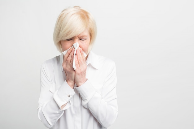 Vieille femme malade éternue dans un mouchoir. On dirait qu'elle a attrapé froid. Elle doit suivre un traitement pour aller mieux. Fermer. Isolé sur blanc