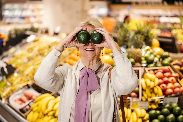 Une vieille femme maladroite joue avec un avocat et fait des grimaces au supermarché