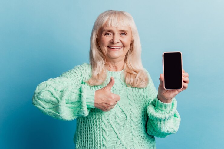 Vieille femme positive tenir l'écran tactile du smartphone afficher le pouce isolé sur fond bleu