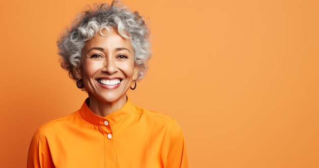 Une vieille femme souriante aux cheveux gris et aux boucles d'oreilles portant une chemise orange avec un col blanc sur un fond orange solide