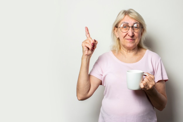 Vieille femme sympathique avec un sourire dans un t-shirt décontracté et des lunettes tient une tasse dans ses mains et pointe son doigt sur un mur lumineux isolé. Visage émotionnel. Faites attention à penser