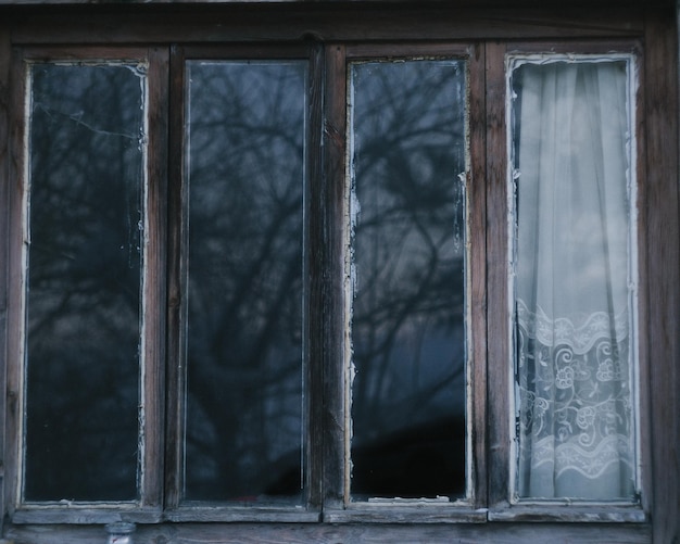 Vieille fenêtre en bois rouillée avec des arbres reflétés sur du verre avec un rideau blanc