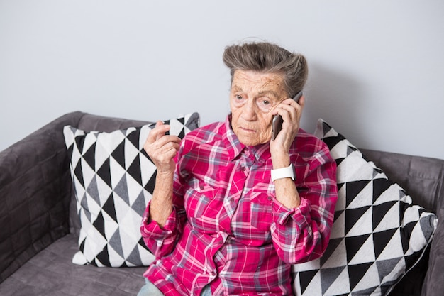 Une vieille grand-mère de femme âgée aux cheveux gris est assise à la maison sur le canapé à l'aide du téléphone portable