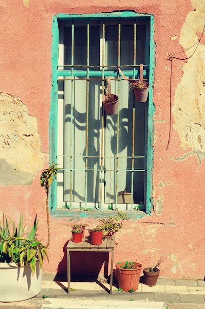 Vieille maison peinte en rose avec des murs fissurés, une fenêtre en bois bleue et des plantes vertes. Concept pop art, style rétro