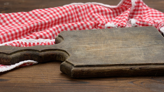 Vieille planche à découper en bois vide et serviette de cuisine pliée en coton rouge et blanc sur fond marron en bois, vue de dessus, espace pour copie