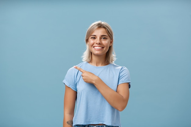 Viens avec moi. Portrait de l'élégante heureuse étudiante aux cheveux blonds en t-shirt à la mode pointant vers le coin supérieur gauche et souriant avec plaisir et bonheur posant