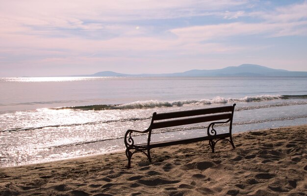 Le vieux banc vide se tient sur la plage sablonneuse et la réflexion du soleil sur l'eau