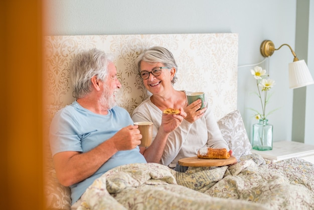 Vieux couple caucasien âgé riant et appréciant le petit déjeuner le matin au lit dans la chambre à coucher à la maison. Couple de personnes âgées mangeant un croissant et buvant du café dans une tasse pour le petit-déjeuner à la maison