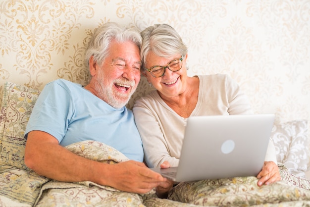 Vieux couple caucasien senior riant et utilisant un ordinateur portable le matin au lit dans la chambre à la maison. Couple de personnes âgées surfant et utilisant les médias sociaux sur un ordinateur portable à la maison.