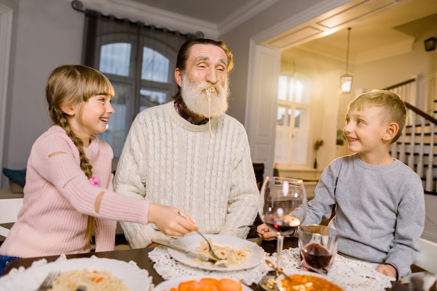 Vieux grand-père avec ses deux petits-enfants assis à la table de la cuisine et mangeant des pâtes. Petite fille et garçon nourrir grand-père avec des pâtes et rire. Moments de vie heureux en famille.