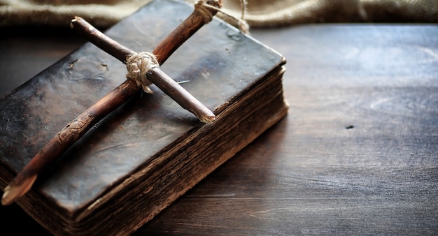 Vieux livre religieux sur une table en bois. Une croix religieuse attachée avec une corde et une toile de jute à côté de la bible. Adoration, péchés et prière.