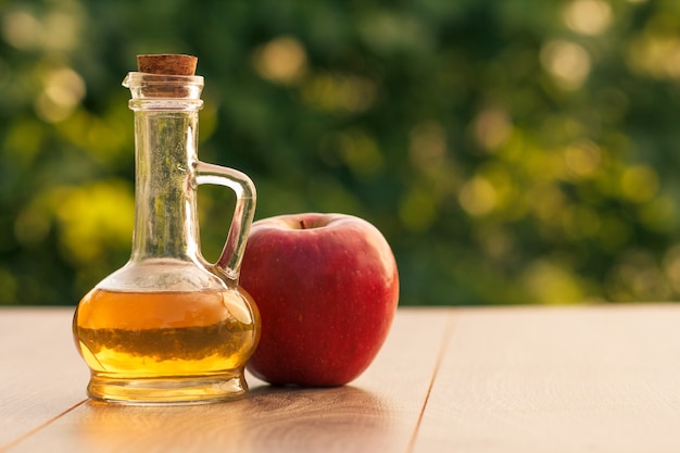 Vinaigre de pomme dans une bouteille en verre avec du liège et pomme rouge fraîche sur des planches de bois avec fond naturel vert flou. Aliments biologiques pour la santé