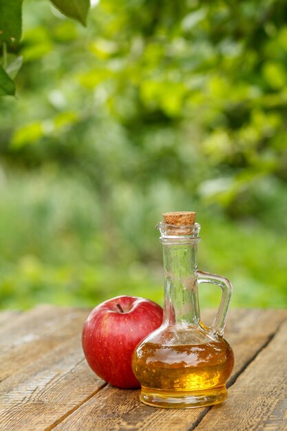 Vinaigre de pomme dans une bouteille en verre avec du liège et pomme rouge fraîche sur de vieilles planches de bois avec fond naturel vert flou. Aliments biologiques pour la santé