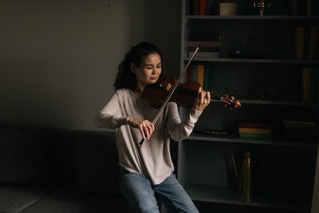 Photo le violoniste de la belle jeune femme joue du violon à la maison, assis sur le canapé