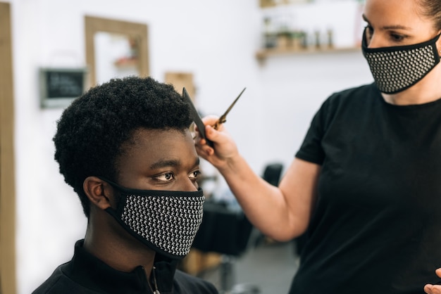Visage d'un black se faisant couper les cheveux dans un salon de coiffure avec un masque noir sur le visage du coronavirus. Le coiffeur porte également un masque. Les cheveux l'ont comme l'afro