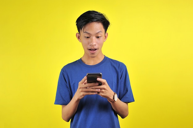 Visage choqué d'un homme asiatique en chemise blanche regardant l'écran du téléphone sur fond jaune.