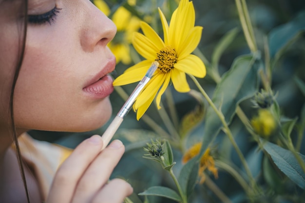 Visage de femme d'été Portrait en gros plan d'une jeune femme séduisante peignant des fleurs jaunes avec une brosse Concept de jardin de fleurs et de printemps Belle femme marchant dans le jardin de fleurs