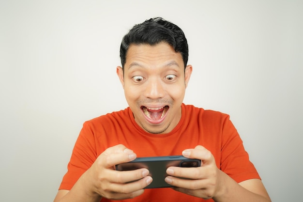 Le visage fou, drôle et excité d'un homme asiatique en T-shirt orange accro au jeu mobile.