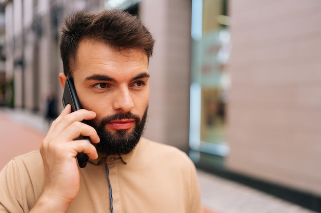Visage en gros plan d'un beau jeune homme barbu marchant dans la rue de la ville et parlant sur un smartphone sur fond de bâtiment
