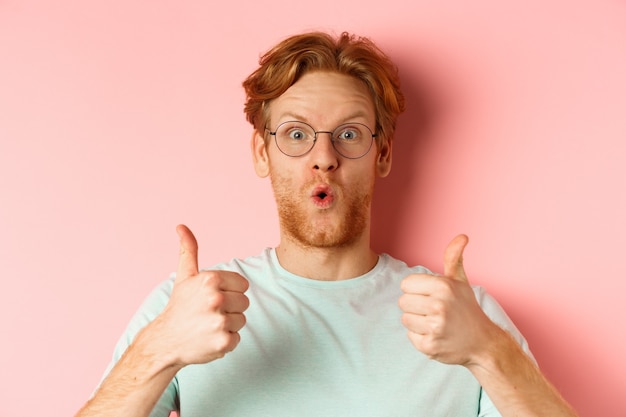 Visage d'heureux homme rousse à lunettes et t-shirt, montrant le pouce levé et l'air excité, approuve et loue une promotion cool, debout sur fond rose.